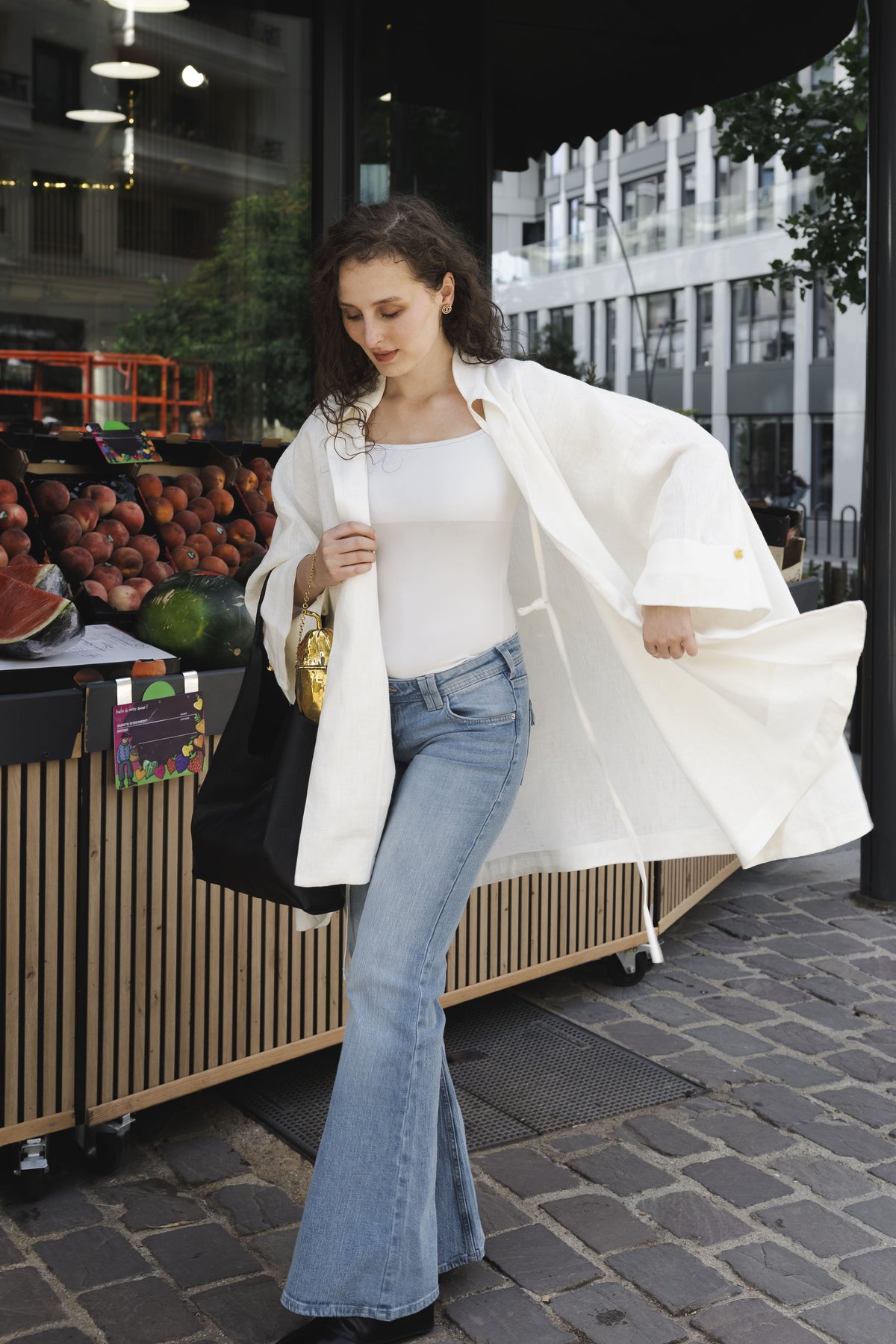 MAATHI Souffle white linen coat worn with blue jeans beside a fruit stand in Paris.