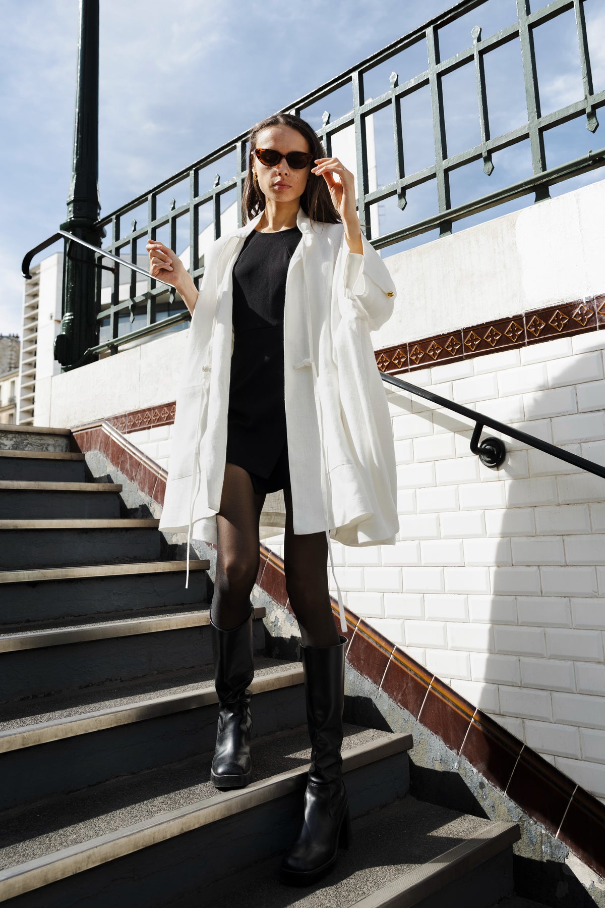 MAATHI Echo white linen coat worn with a black dress, sunglasses and boots on Paris metro stairs.