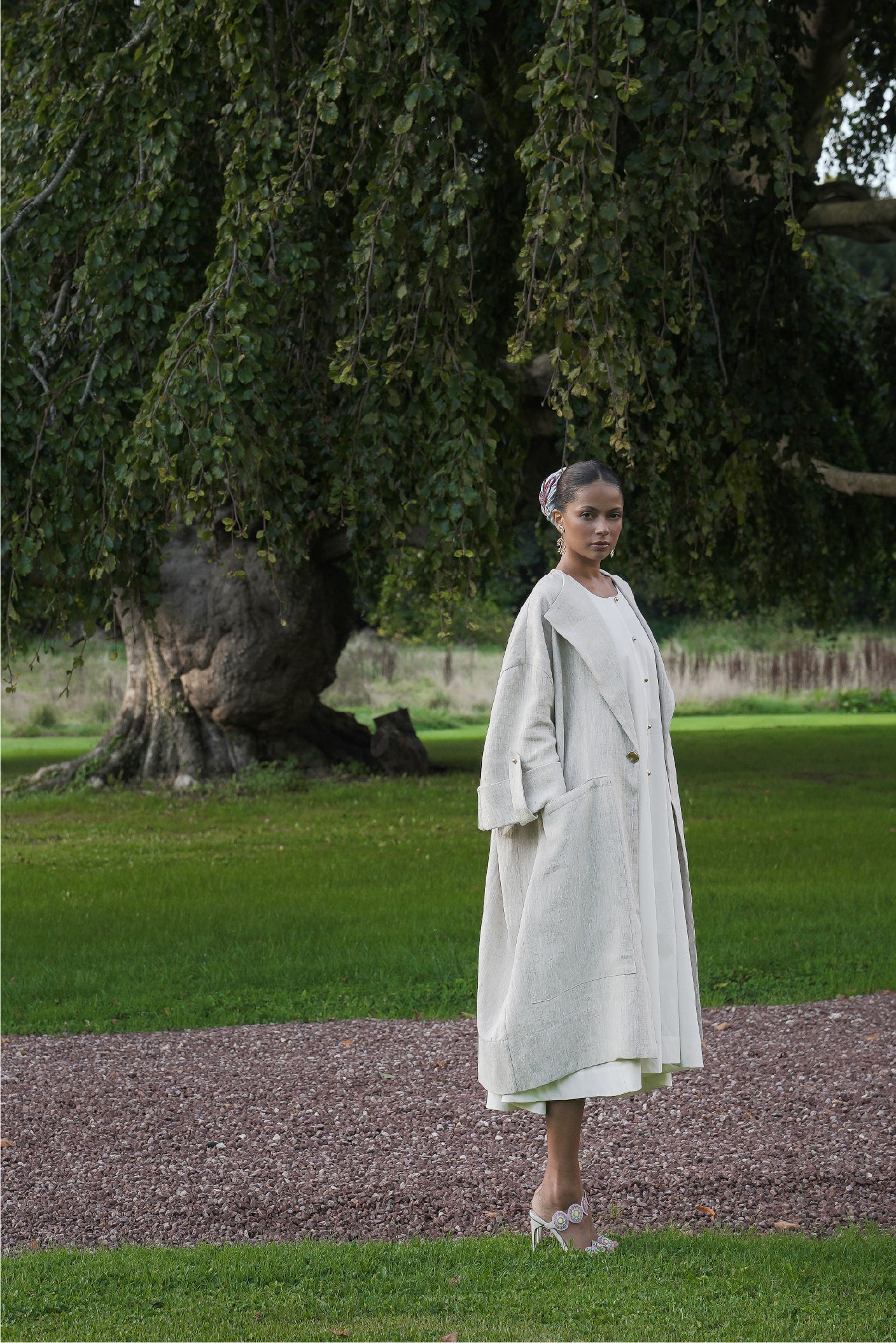 Model wearing the Echo linen coat by MAATHI Paris in a poetic garden editorial setting beneath a large tree