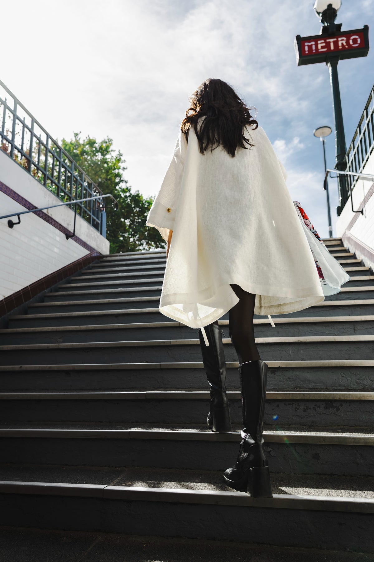 Back view of the MAATHI souffle white linen coat worn with black boots on Paris metro stairs. |
