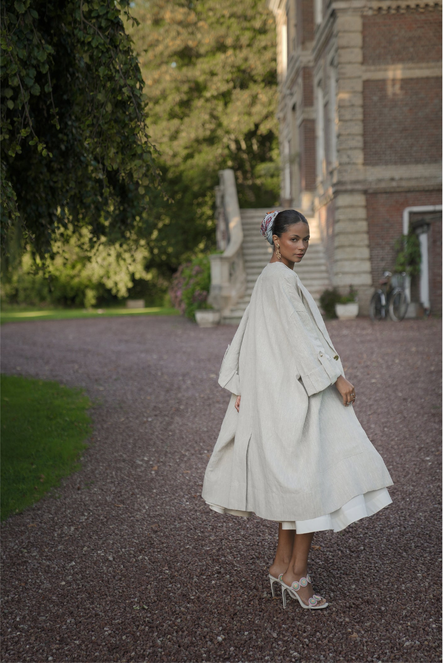 Model wearing the Echo Linen Coat with the Tide Silk Scarf tied in her hair, standing on a garden path outside a château