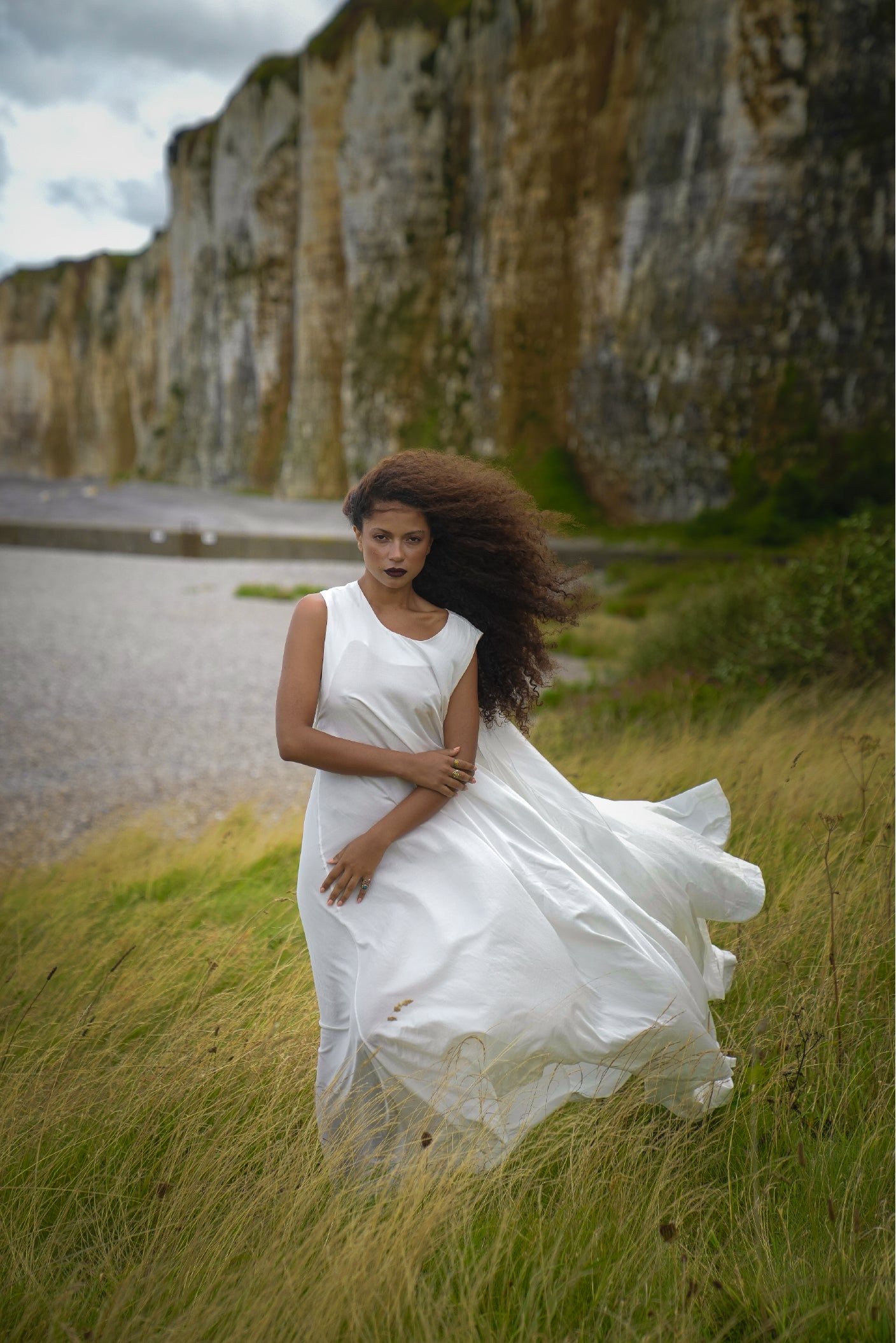 Model wearing the Moonwake Dress in flowing white fabric, standing in tall grass beside dramatic coastal cliffs