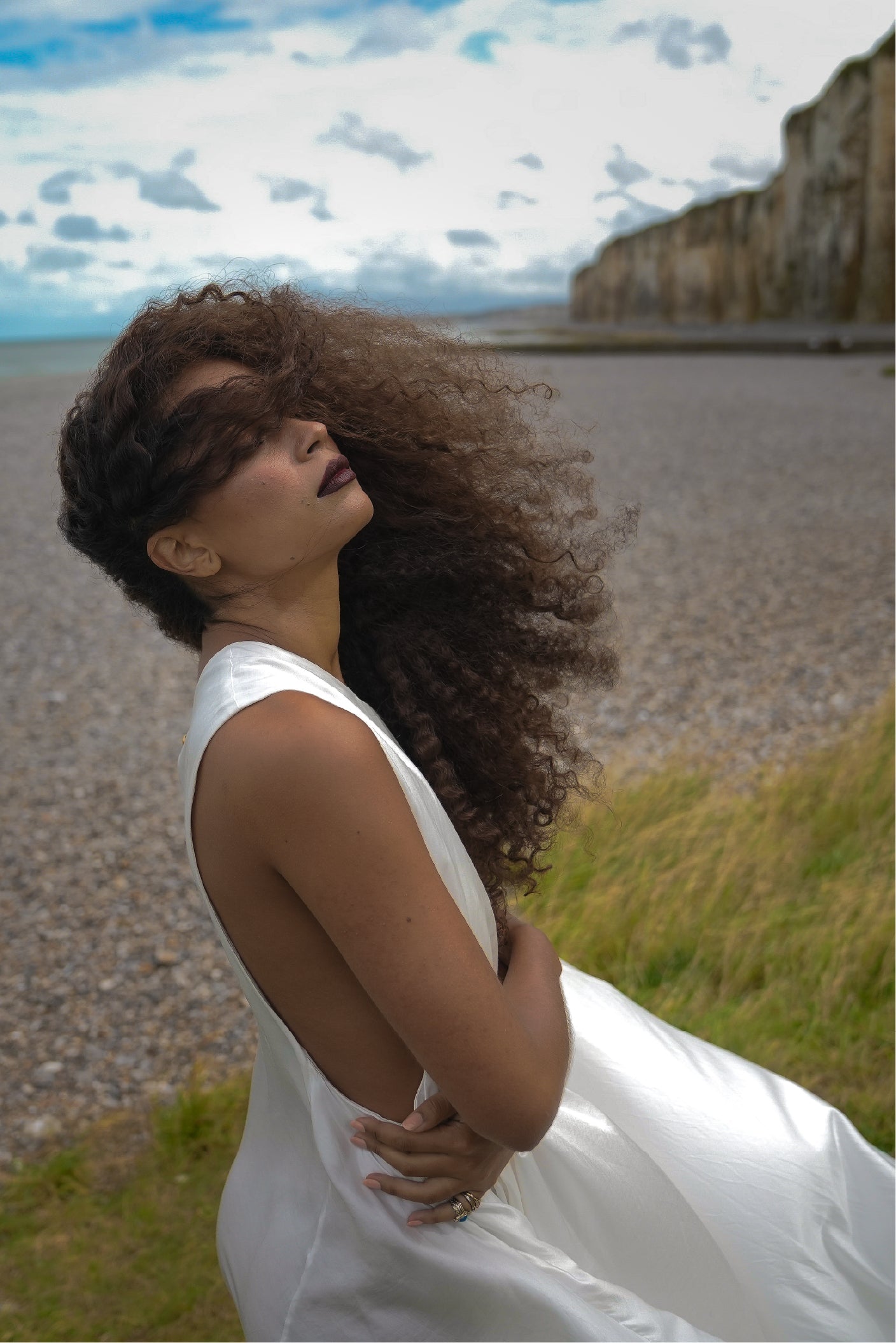 Close portrait of a model wearing the Moonwake Dress beside coastal cliffs, with windblown curls and flowing white fabric.